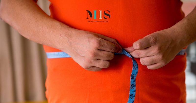 Man in orange shirt measures his waist to track his progress after weight loss surgery at MIIS Weight Loss Institute in St. Pete, FL