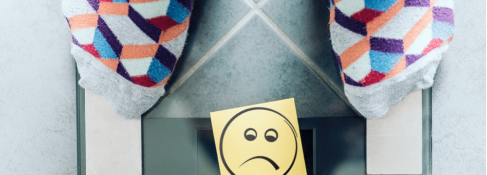 Feet on bathroom scale with frowning face on note over the numbers noting a stall in weight loss 